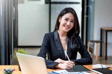 Wall Mural - Beautiful young Asian businesswoman holding a pen with a tablet accounting work. Looking at the camera.