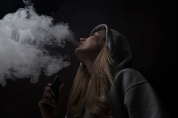 Wall Mural - Portrait of beautiful blonde girl in gray hoodie smokes vape isolated on black studio background, cloud of steam smoke, mini hookah