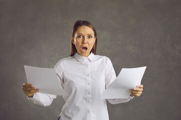 Portrait business woman holding expensive utility bill or office paper document with accounting mistakes looking at camera with shocked surprised astonished face expression isolated on grey background