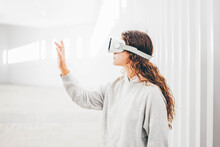 Curly Haired Young Woman In Virtual Reality Glasses Moves Hands Touching Online Object And Looks Around Standing In White Futurisctic Tunnel