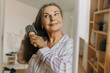 © shurkin_son - Indoor portrait of attractive mature Caucasian woman in striped pajama brushing hair while taking care of herself, standing in front of mirror, after waking up in early morning, having make-up