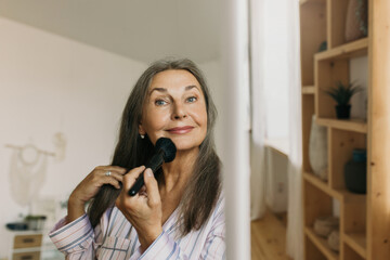 Wall Mural - Beautiful 60-years-old lady in casual clothes doing makeup at home standing in front of mirror at her bedroom or hallway, preparing for going out with her friends, using face brush to apply blusher