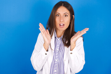 Wall Mural - Surprised terrified young european doctor girl wearing white medical gown ​on white background Gestures with uncertainty, stares at camera, puzzled as doesn't know answer on tricky question, 