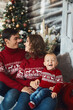 © innarevyako - Funny little kid in an ugly sweater posing with his parents over Christmas interior. Young parents and their little son in Christmas clothes