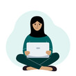 A woman in a black headscarf sits and works at a laptop. Muslim. Remote work or communication over the Internet.