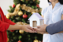 Woman Realtor Hands Toy House To Couple Against Background Of New Year Tree