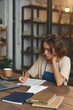 © pressmaster - Busy female owner sitting at wooden table and making notes in papers while working with finances in pottery shop
