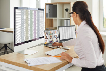 Busy young female employee sitting at her office table in front of multiple computers, working with financial business spreadsheets and having a video conference call with colleagues at the same time