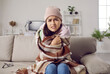 © Studio Romantic - It's so cold in house in winter. Woman freezes in wintertime due to problems with central heating. Young girl wearing warm hat and wrapped in wool blanket sitting on sofa at home and looking at camera