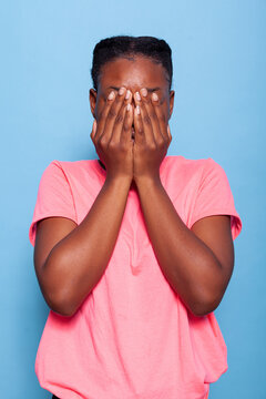 Portrait of anxious emotional african american student covering face with her hands during photo shotting having social anxiety standing in studio with blue background. Terrified introvert model