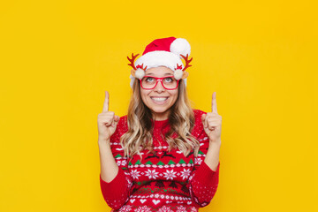 Wall Mural - A young smiling blonde woman in glasses with deer antlers, a red deer sweater and Christmas Santa hat points up to empty copy space for greeting text with fingers isolated on color yellow background