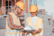 © Migue Suarez - Equipo joven de arquitectos de una constructora para una inmobiliaria trabajando en equipo con cascos mientras revisan los planos de la construcción de obra