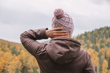 Wall Mural - A female traveler stands on the top of the mountain, admiring the magnificent landscape of the autumn forest from above. Selective focus. Close-up. Eco tourism. People from behind.