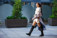 Beautiful sexy fashion model. Lady walks city street passes by. Young woman in coat, black knee high boots. Glamour girl, long bare leg. Elegant dark long hair, pretty face, looking in camera, blurred
