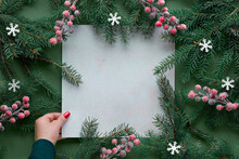 Green Red Christmas Background With Square Copy-space, Place For Text. Natural Xmas Twigs Decorated With Frosted Berries And Paper Snowflakes. Flat Lay. Top View On Off White Paper In Female Hand.