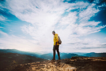 Wall Mural - Young hiker man standing on top of cliff in summer mountains at sunset and enjoying view of nature