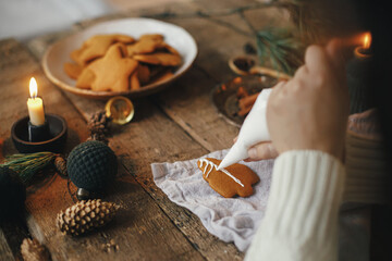 Hands decorating gingerbread cookie christmas tree with frosting on rustic table with napkin, candle, spices, decorations. Atmospheric moody image. Making traditional christmas gingerbread cookies