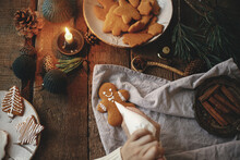Hands decorating christmas gingerbread man cookie with icing on rustic table with napkin, spices, candle, decorations. Flat lay. Moody image. Woman making stylish christmas gingerbread cookies