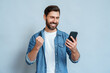 Happy delighted satisfied adult man looking at mobile phone screen gesturing yes winning pose, isolated background. Overjoyed excited joyful guy make winner gesture read message about happy good news