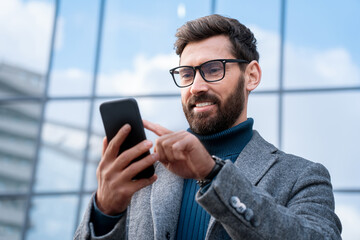 Wall Mural - smiling businessman using smartphone standing outside office in urban street