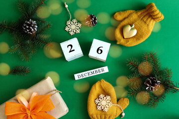 Calendar for December 26 : the name of the month in English, cubes with the number 26, yellow mittens, a gift with a yellow bow, spruce branches on a green background, top view, bokeh