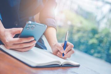 Wall Mural - Business woman hand writing on a notepad with a pen and she is holding a cell phone in the office.