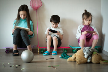 Wall Mural - Three funny serious children sitting on skateboards and playing mobile games on gadgets at home