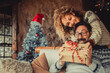 © simona - Overjoyed man receive christmas gift from his wife. Happy people enjoy and celebrate christmas eve at home. Xmas tree in background. Winter leisure couple lifestyle indoor