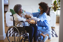 African American Female Doctor With Face Mask Treating Senior Female Patient On Wheelchair At Home