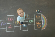 Little Boy And Colorful Hopscotch Drawn With Chalk On Asphalt Outdoors, Top View. Happy Childhood
