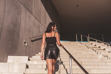 Dark-haired Woman Posing In A Black Dress On The Concrete Stairs. The Woman Is Wearing Black Sunglasses. It's A Beautiful Sunny Day.