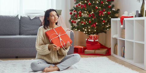 Young adult woman holding present in a wrapped red gift box, sitting in the living room at home in front of Christmas tree background. Smiling girl spending her winter holidays indoors