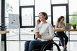 © LIGHTFIELD STUDIOS - smiling businesswoman in wheelchair holding coffee to go and talking on cellphone near blurred african american colleague