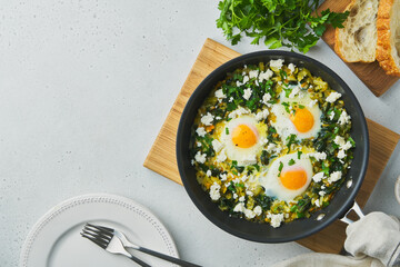 Wall Mural - Delicious mediterranean green shakshouka fried with eggs, spinach, celery, salty sheep milk cheese, pepper and ciabatta. Served in pan on wooden cutting board. Copy space, horizontal