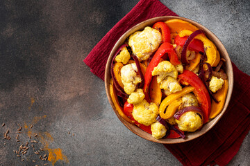 Wall Mural - Baked spicy vegetables in a bowl on a dark background top view. Pepper, sweet potato, cauliflower and onion baked with cumin, turmeric, curry and ginger. Indian food, vegan food. Copy space