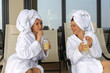 Two happy attractive brunette female friends in white bathrobes and white towel on head have fun on sunbed near hotel swimming pool.