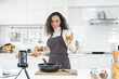 © LOVEis - Latin women live streaming online with a smartphone while cooking in the kitchen at home.