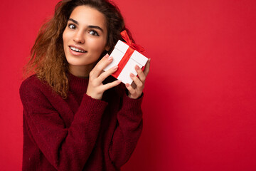 Closeup photo of attractive positive smiling young brunette curly woman isolated over red background wall wearing red sweater holding gift box looking at camera and dreaming