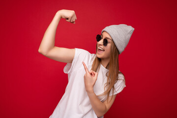 Wall Mural - Portrait of young emotional positive happy beautiful dark blonde woman with sincere emotions wearing casual white t-shirt with empty space for mockup gray hat and sunglasses isolated over red