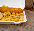 Close and selective focus on a polystyrene carton comprising a battered jumbo sausage and traditional chip shop chips cooked in beef dripping on an outdoor wooden table