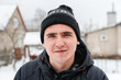 Close-up portrait of young man in warm hat outside on rural winter snowy house background. Happy millennial smiling outdoor, cold weather. Caucasian guy 20-25 years. Face with acne or herpes