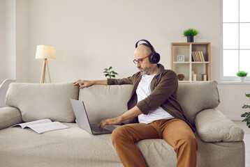 Canvas Print - Adult relaxed man listening to music on headphones while working on laptop at home. Middle-aged Caucasian man sits next to laptop and notebook on comfortable sofa in cozy living room.