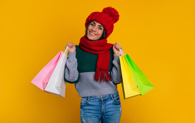 Wall Mural - Close up photo of attractive young woman in red winter hat and scarf with colorful paper bags for purchase and gifts isolated on yellow background