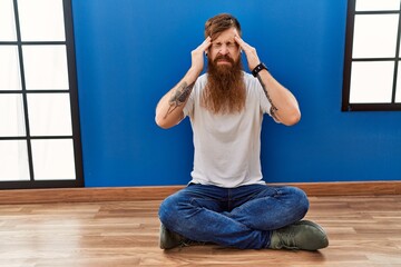 Poster - Redhead man with long beard sitting on the floor at empty room with hand on head for pain in head because stress. suffering migraine.