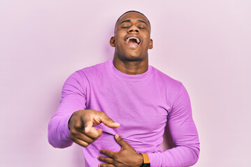 Young black man wearing casual pink sweater laughing at you, pointing finger to the camera with hand over body, shame expression