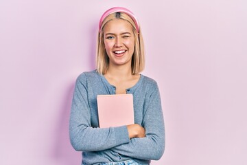 Canvas Print - Beautiful blonde woman holding book winking looking at the camera with sexy expression, cheerful and happy face.