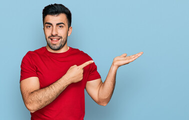 Wall Mural - Hispanic man with beard wearing casual red t shirt amazed and smiling to the camera while presenting with hand and pointing with finger.