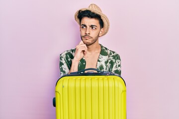 Wall Mural - Young hispanic man wearing summer style and holding cabin bag thinking concentrated about doubt with finger on chin and looking up wondering