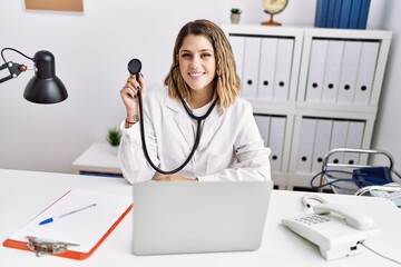 Sticker - Young hispanic woman wearing doctor uniform holding stethoscope at medical clinic looking positive and happy standing and smiling with a confident smile showing teeth