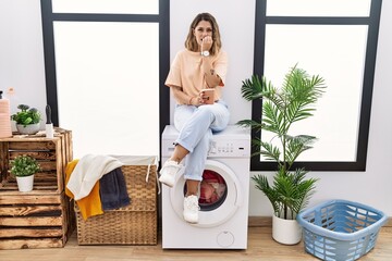 Canvas Print - Young hispanic woman drinking coffee waiting for washing machine at laundry room looking stressed and nervous with hands on mouth biting nails. anxiety problem.